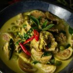 Thai green beef curry with eggplant garnished with Thai basil leaves and sliced red chilies in a blue bowl on a black surface