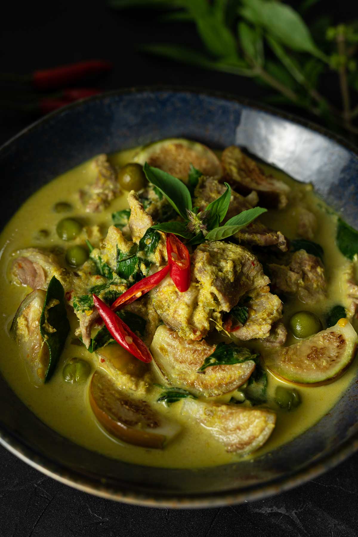 Thai green beef curry with eggplant garnished with Thai basil leaves and sliced red chilies in a blue bowl on a black surface